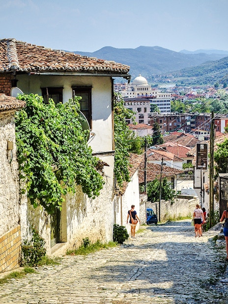 Guests walking along cobblestone street in Berat Old Town, Albania, with historic buildings and distant hills.