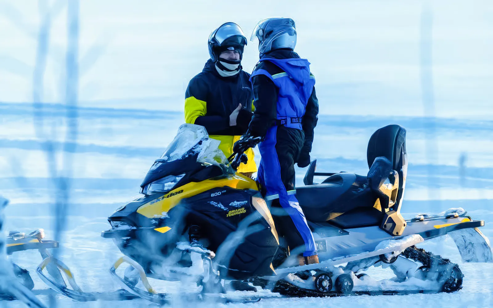 Two people in helmets standing by a snowmobile on a snowy landscape in Tromso.