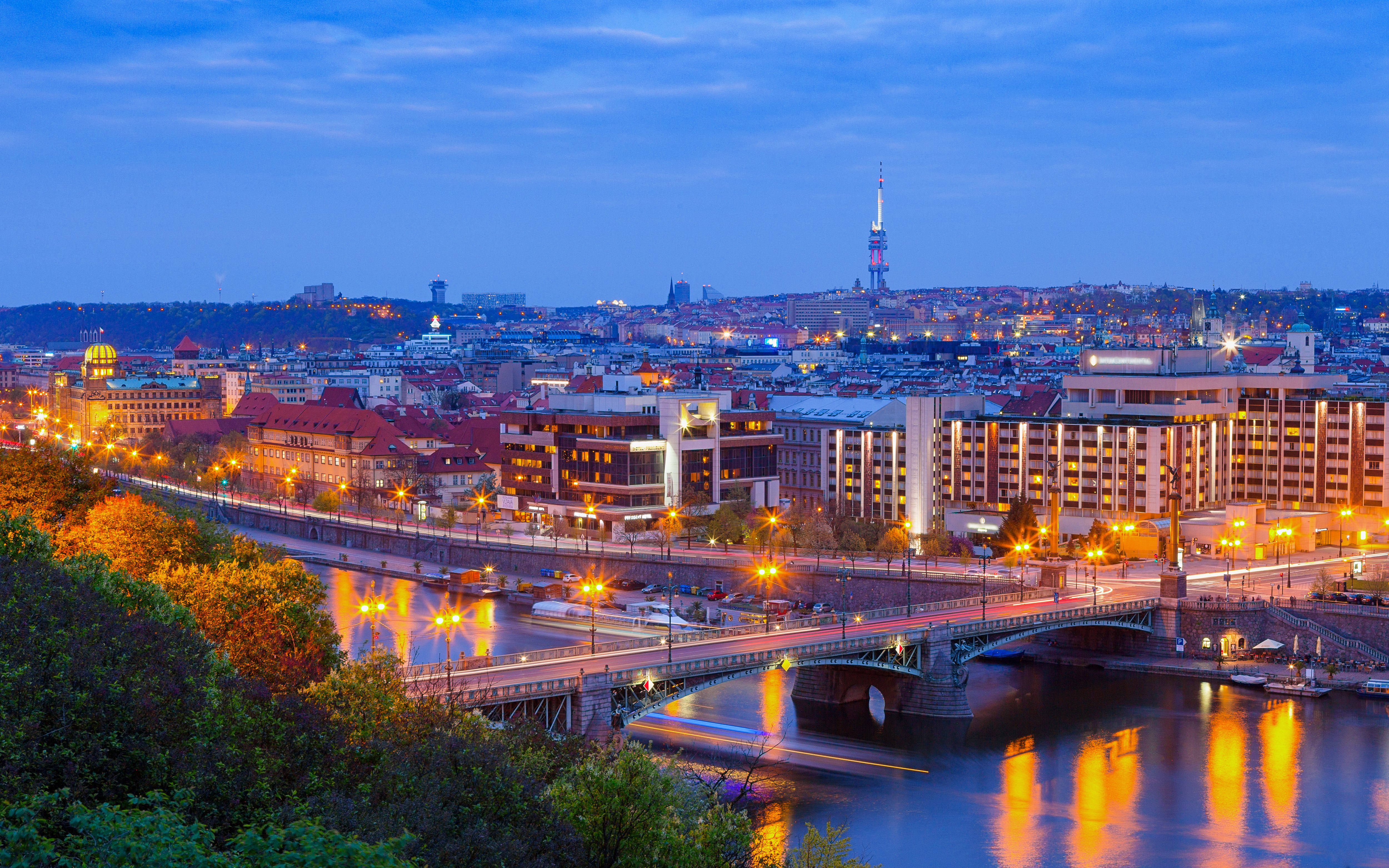 Cechuv Bridge illuminated at night with Prague cityscape in the background.