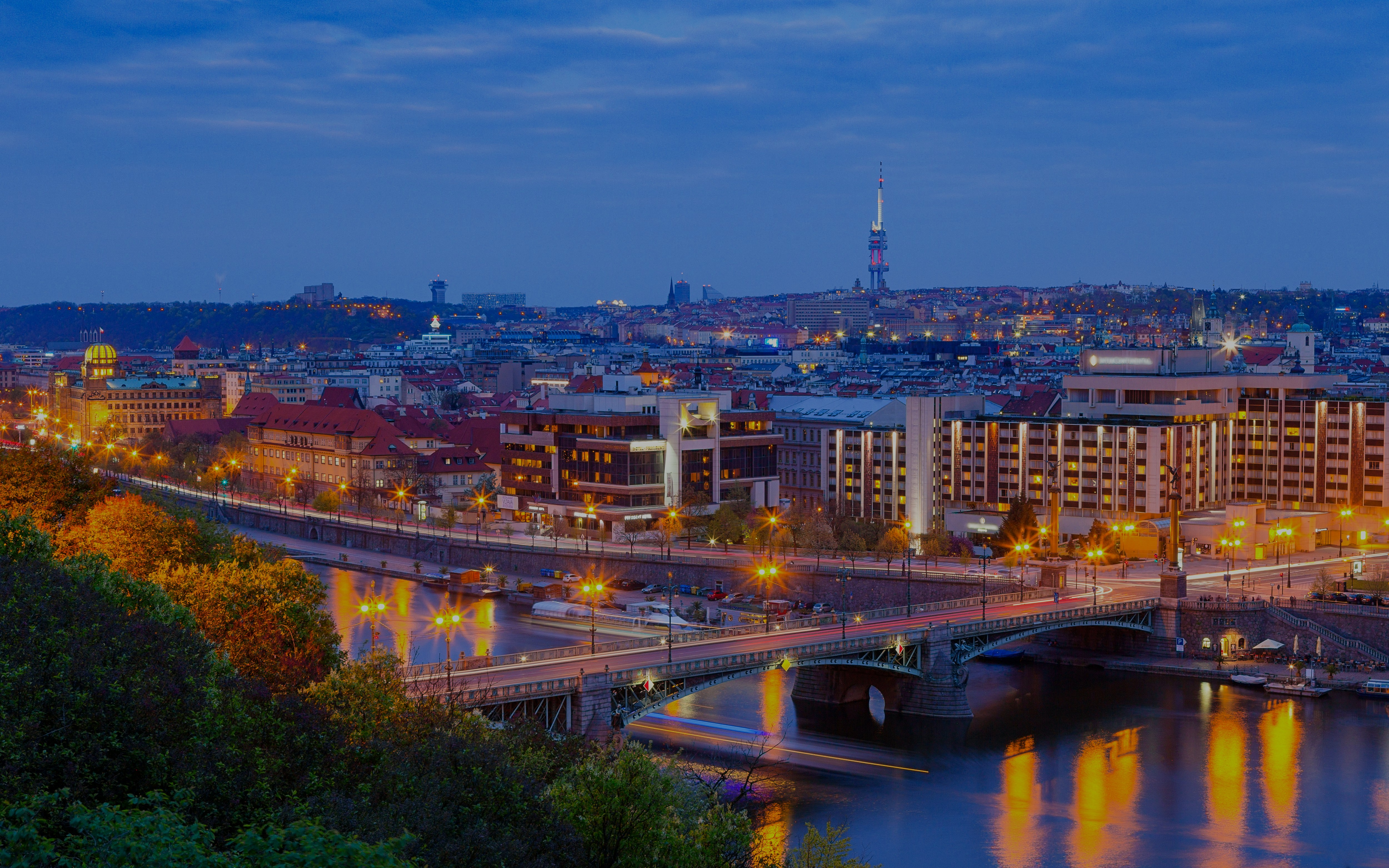 Cechuv Bridge illuminated at night with Prague cityscape in the background.