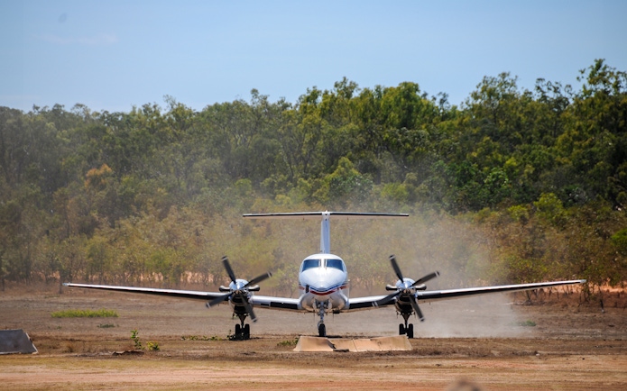 Small aircraft landing on a dirt runway surrounded by trees, part of Royal Flying Doctor Service tour.