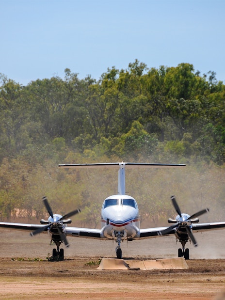 Small aircraft landing on a dirt runway surrounded by trees, part of Royal Flying Doctor Service tour.