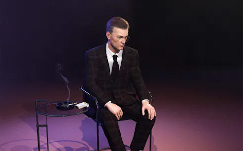 Man in a suit sitting on a chair next to a table with a cigarette and ashtray, Ed Atkins, The Worm, 2021.