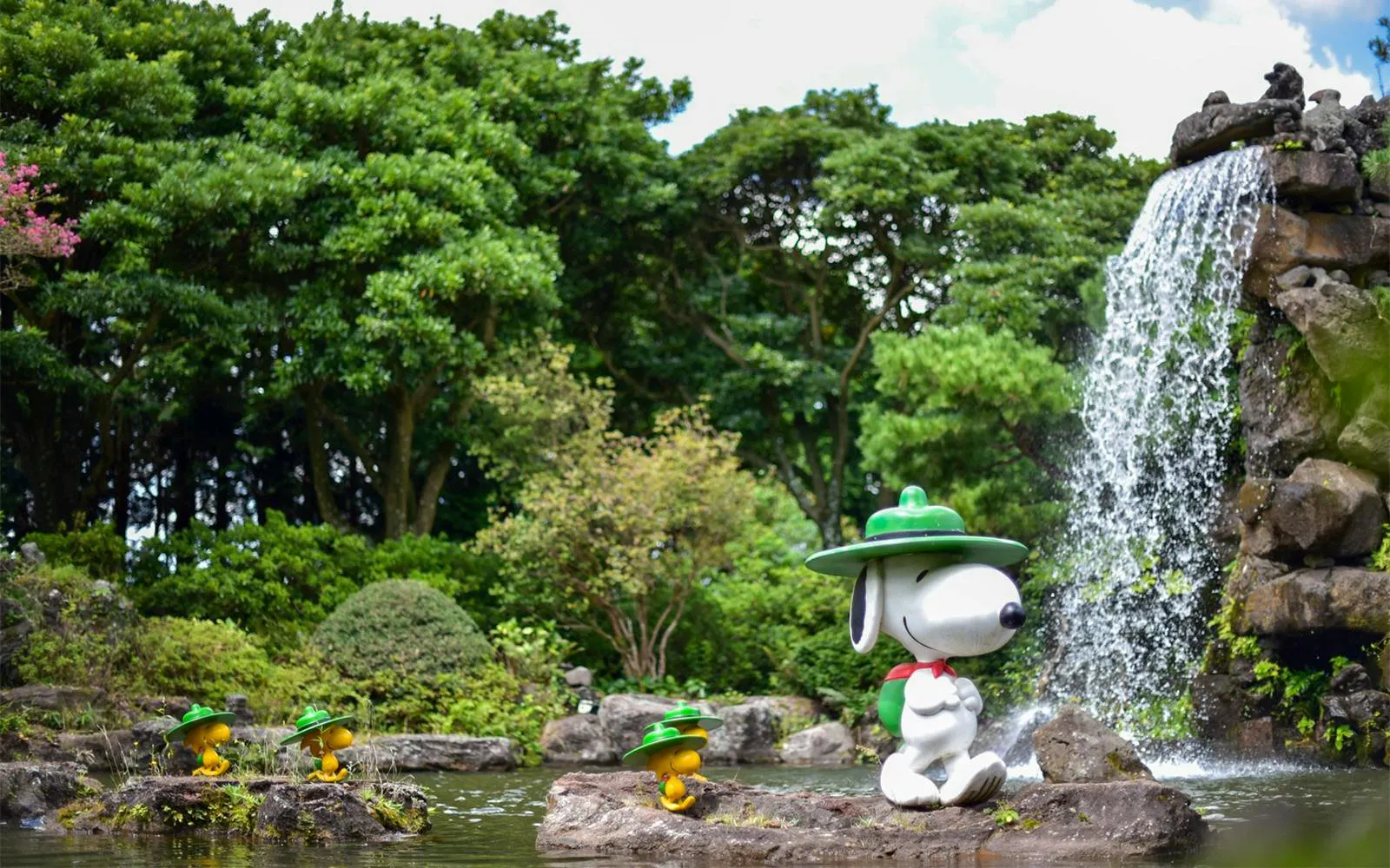 Snoopy statue by waterfall in Snoopy Garden, Jeju Island, surrounded by lush greenery.