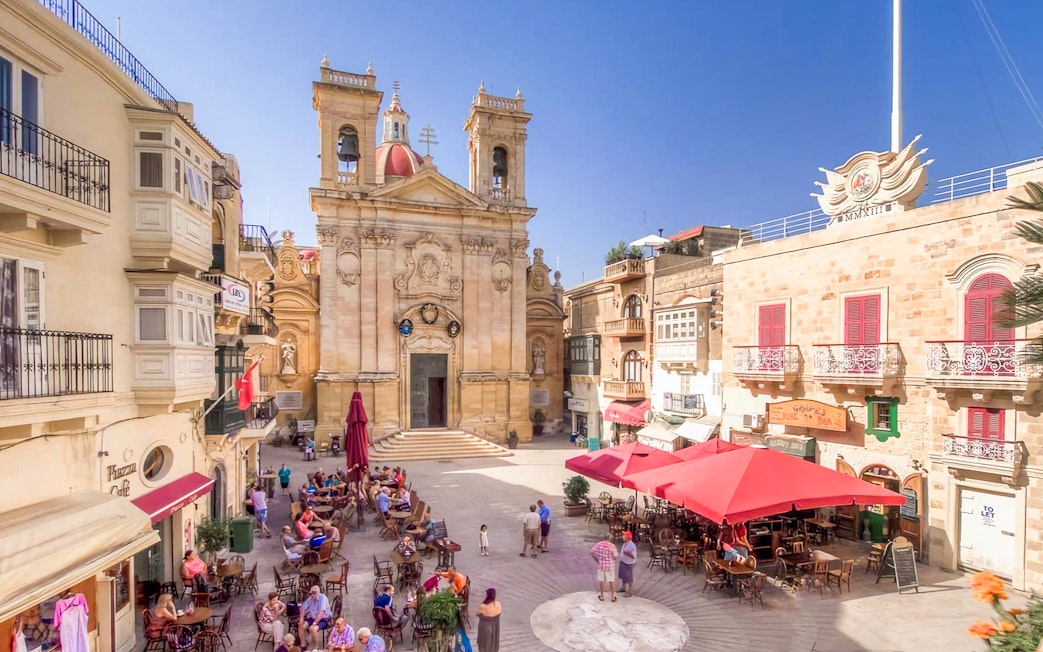 Square in Gozo with people dining at outdoor cafes, featuring a historic church facade.