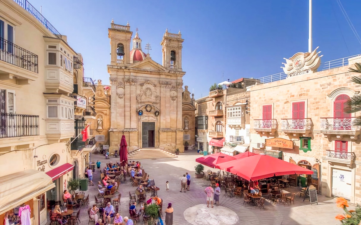 Square in Gozo with people dining at outdoor cafes, featuring a historic church facade.