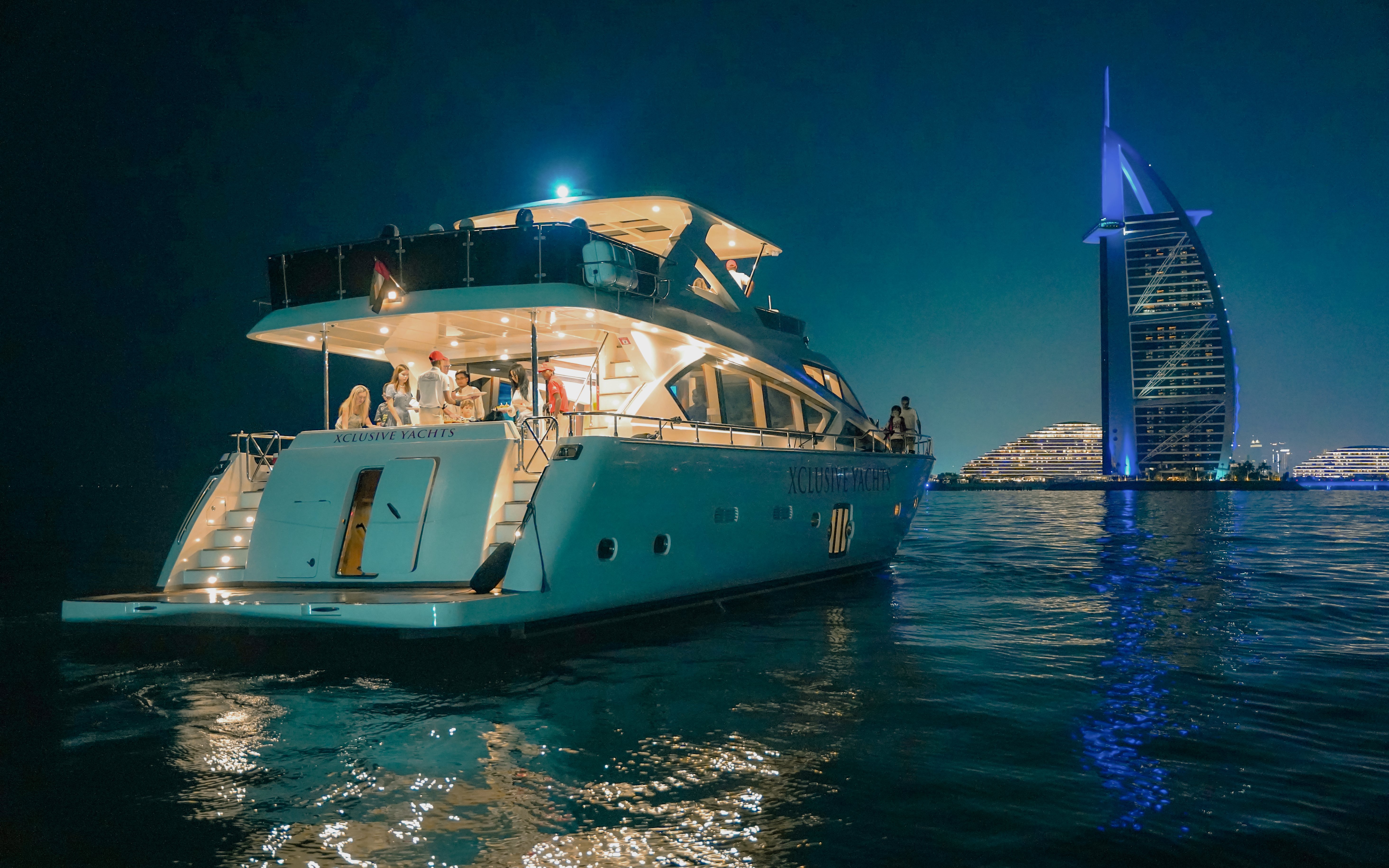 Luxury yacht with guests near Burj Al Arab, Dubai Harbour at night.