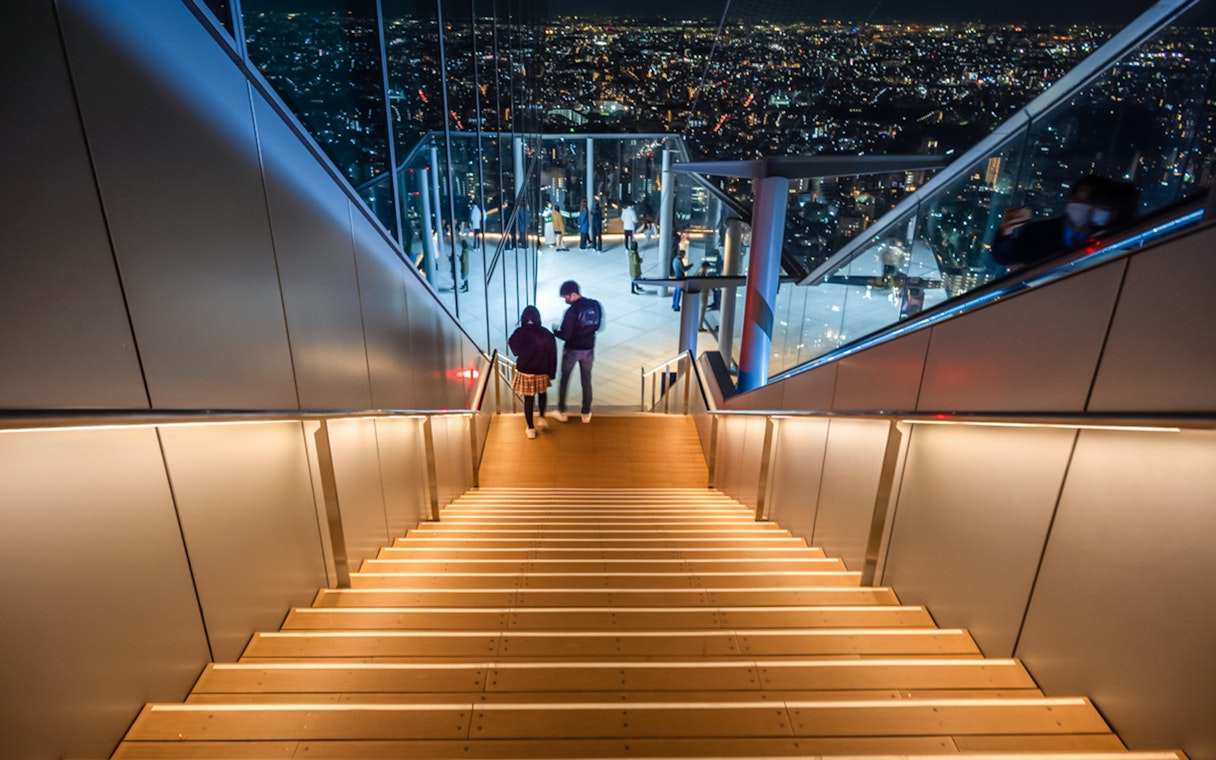 Visitors descending stairs at Shibuya Sky Deck with Tokyo cityscape view.