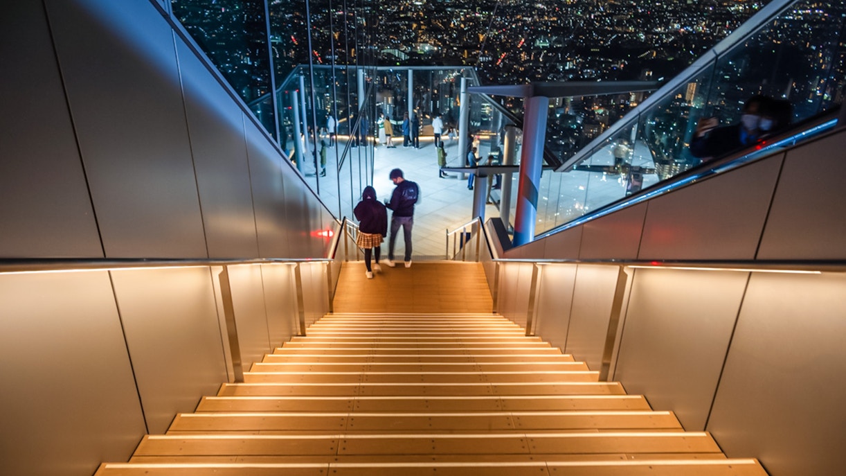 Visitors descending stairs at Shibuya Sky Deck with Tokyo cityscape view.