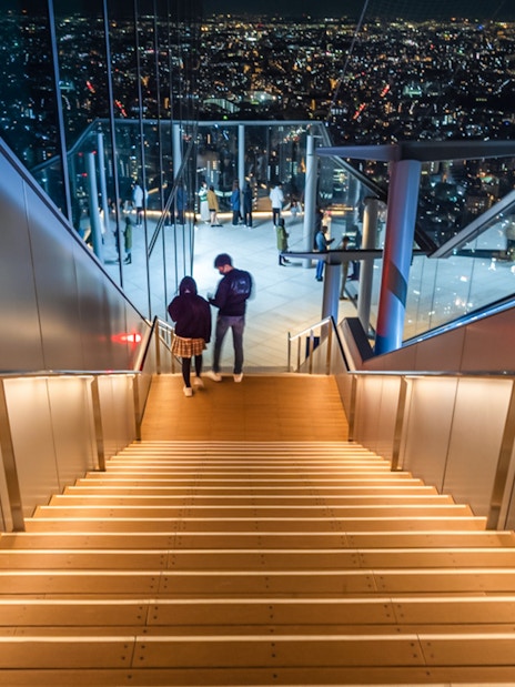 Visitors descending stairs at Shibuya Sky Deck with Tokyo cityscape view.