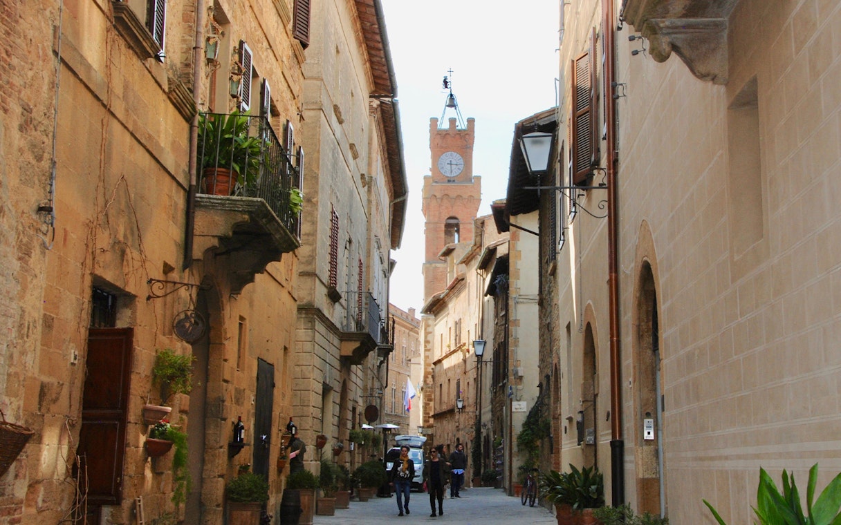 Narrow street in Tuscany with historic buildings and a clock tower in the distance.