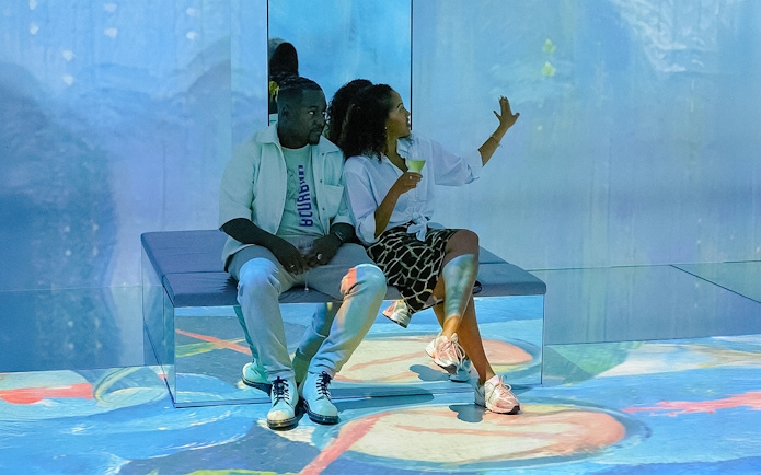 Visitors exploring exhibit at Frameless Lates, seated on a bench with colorful projections.
