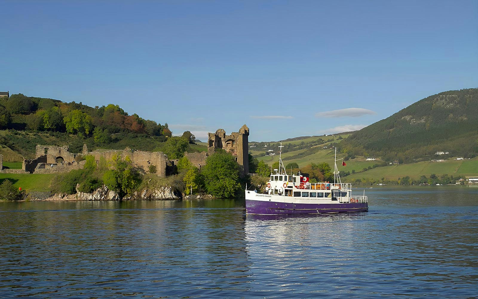 Boat cruising near Urquhart Castle on Loch Ness, Scotland, with green hills in the background.