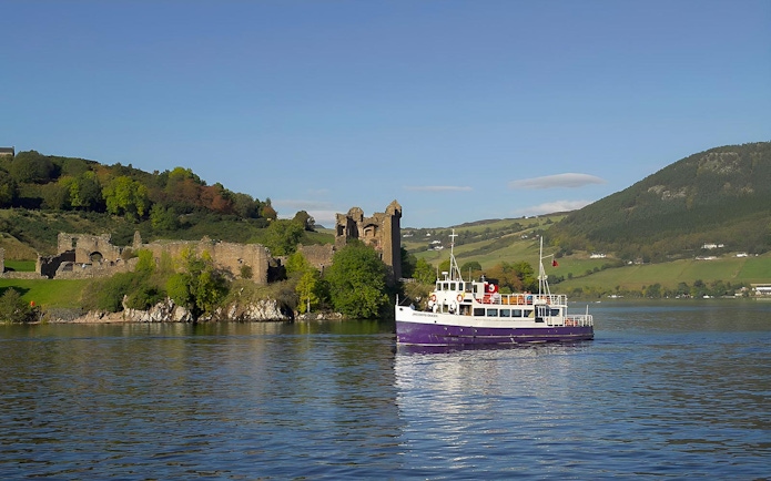 Boat cruising near Urquhart Castle on Loch Ness, Scotland, with green hills in the background.