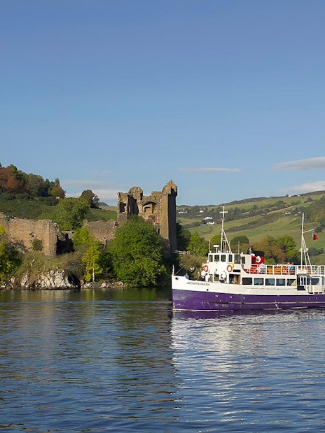 Boat cruising near Urquhart Castle on Loch Ness, Scotland, with green hills in the background.