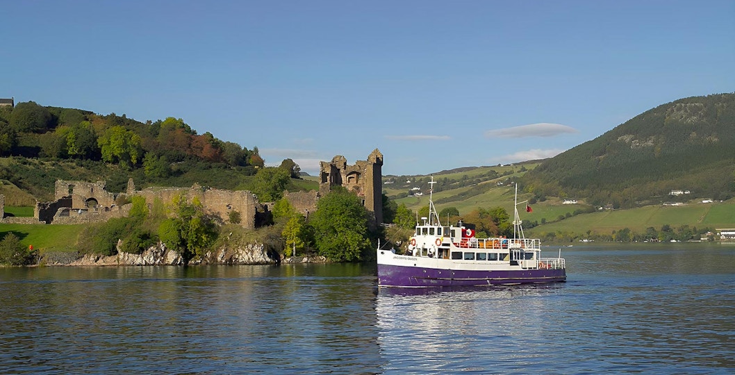 Boat cruising near Urquhart Castle on Loch Ness, Scotland, with green hills in the background.