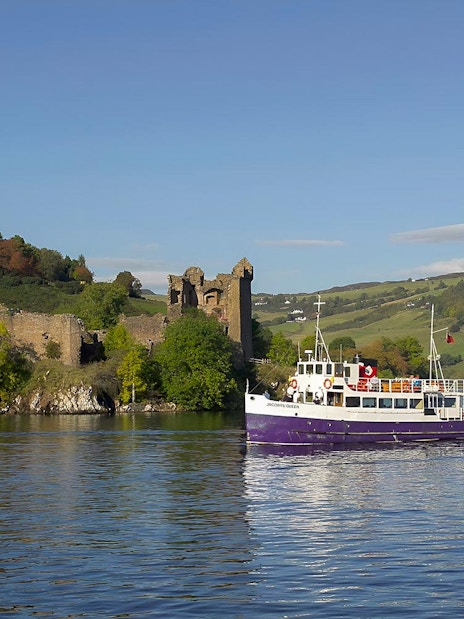 Boat cruising near Urquhart Castle on Loch Ness, Scotland, with green hills in the background.