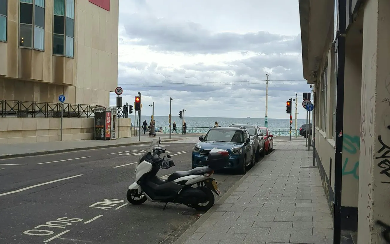 Street view near Brighton seafront with parked vehicles and pedestrians by the ocean.