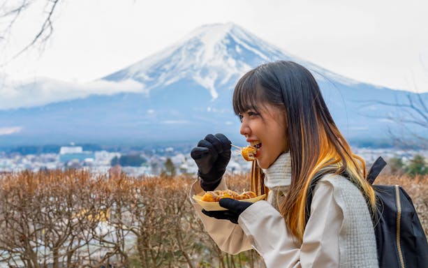 Woman enjoying Takoyaki at Lake Kawaguchi with Mount Fuji in the background, Tokyo tour.