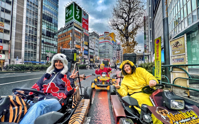 Go-kart riders in costumes driving through Shibuya, Tokyo with cityscape backdrop.