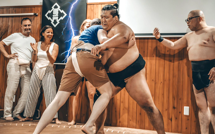 Participant wrestling sumo wrestler in ring, surrounded by cheering spectators and referee.