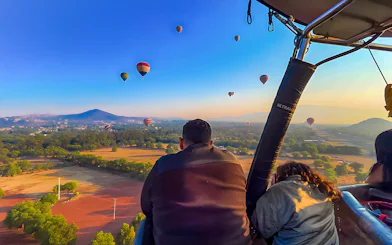 Passengers in a hot air balloon over Teotihuacan with distant balloons and landscape.