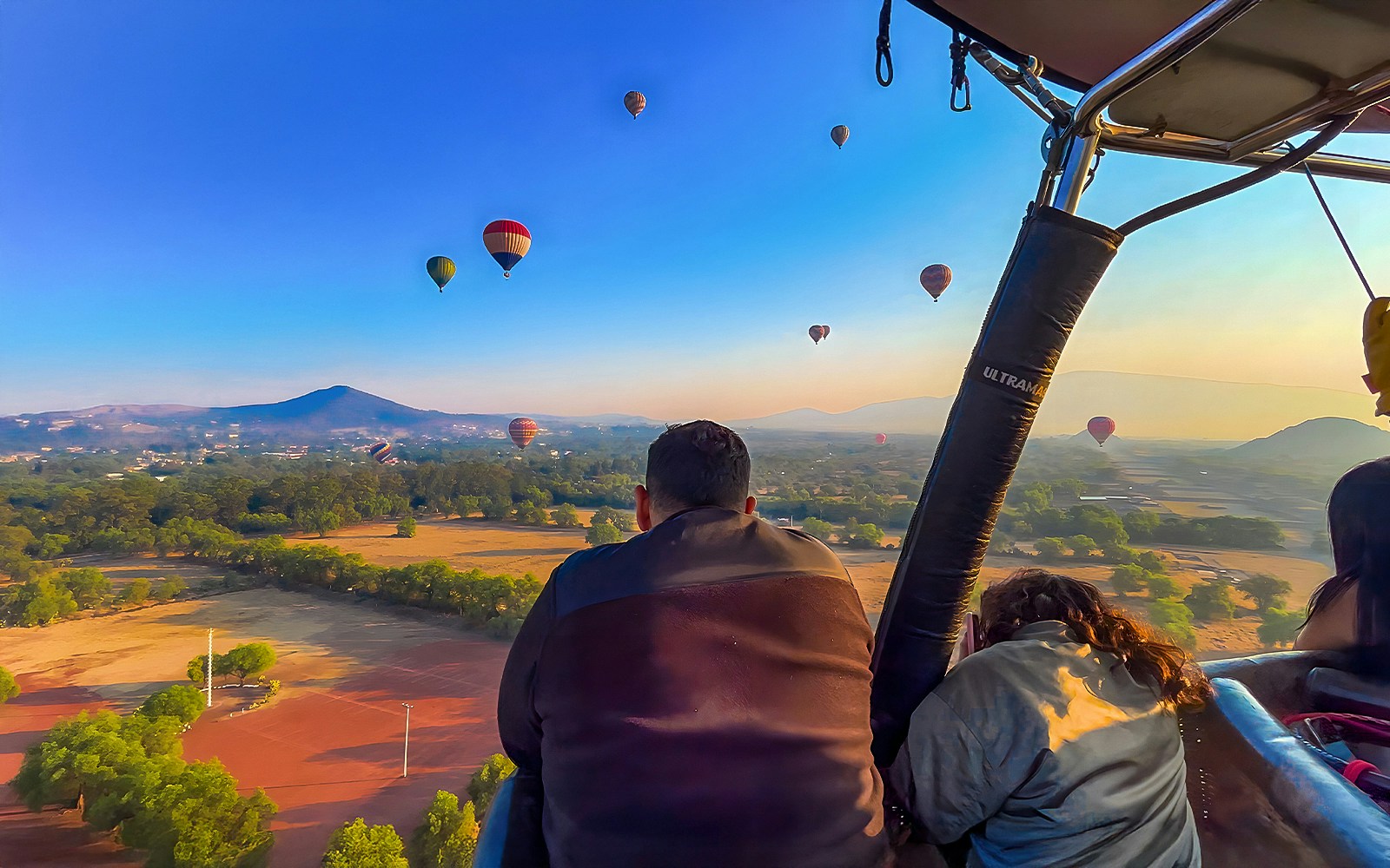 Passengers in a hot air balloon over Teotihuacan with distant balloons and landscape.
