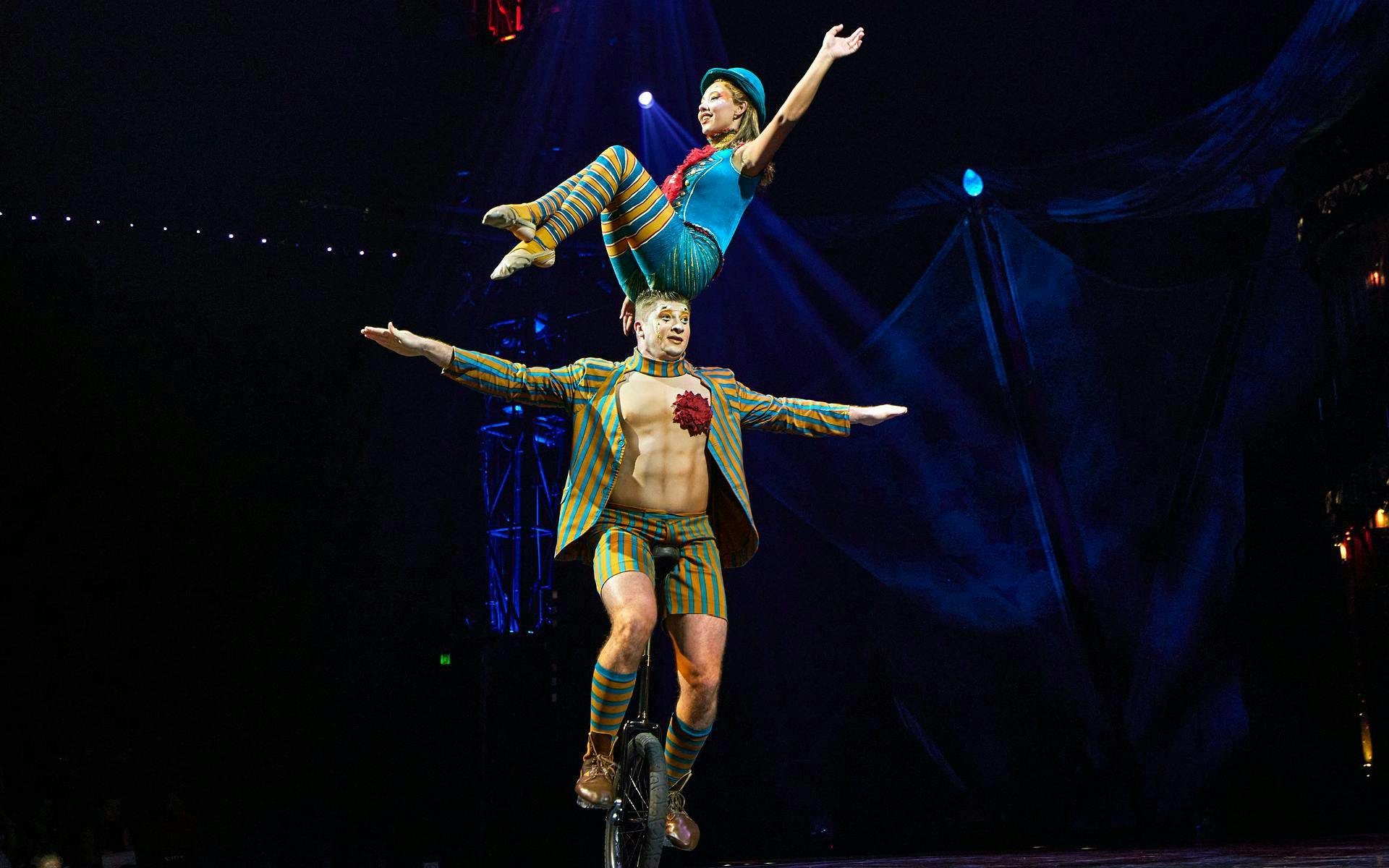 Performer balancing on unicycle with acrobat on shoulders during Kooza stage show.