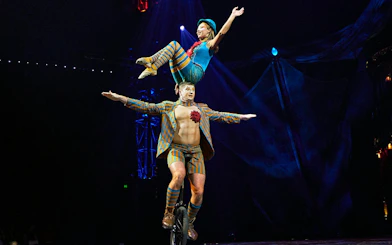 Performer balancing on unicycle with acrobat on shoulders during Kooza stage show.