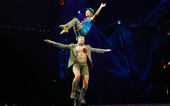 Performer balancing on unicycle with acrobat on shoulders during Kooza stage show.
