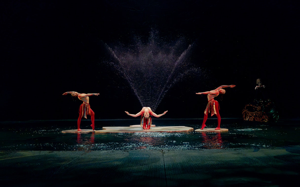 Performers in a water-themed acrobatic show with fountains in the background.