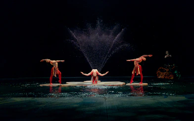 Performers in a water-themed acrobatic show with fountains in the background.