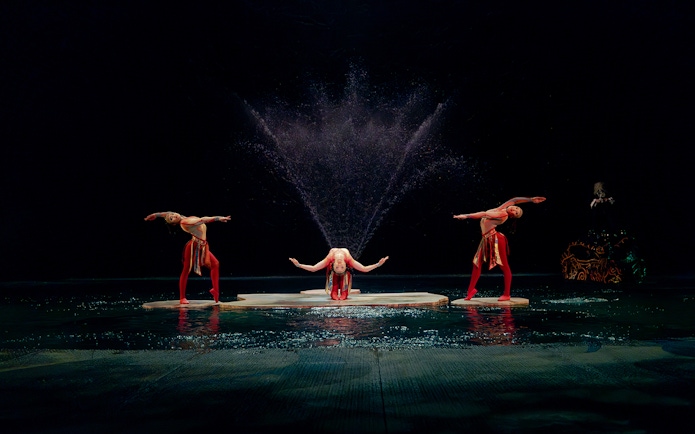 Performers in a water-themed acrobatic show with fountains in the background.