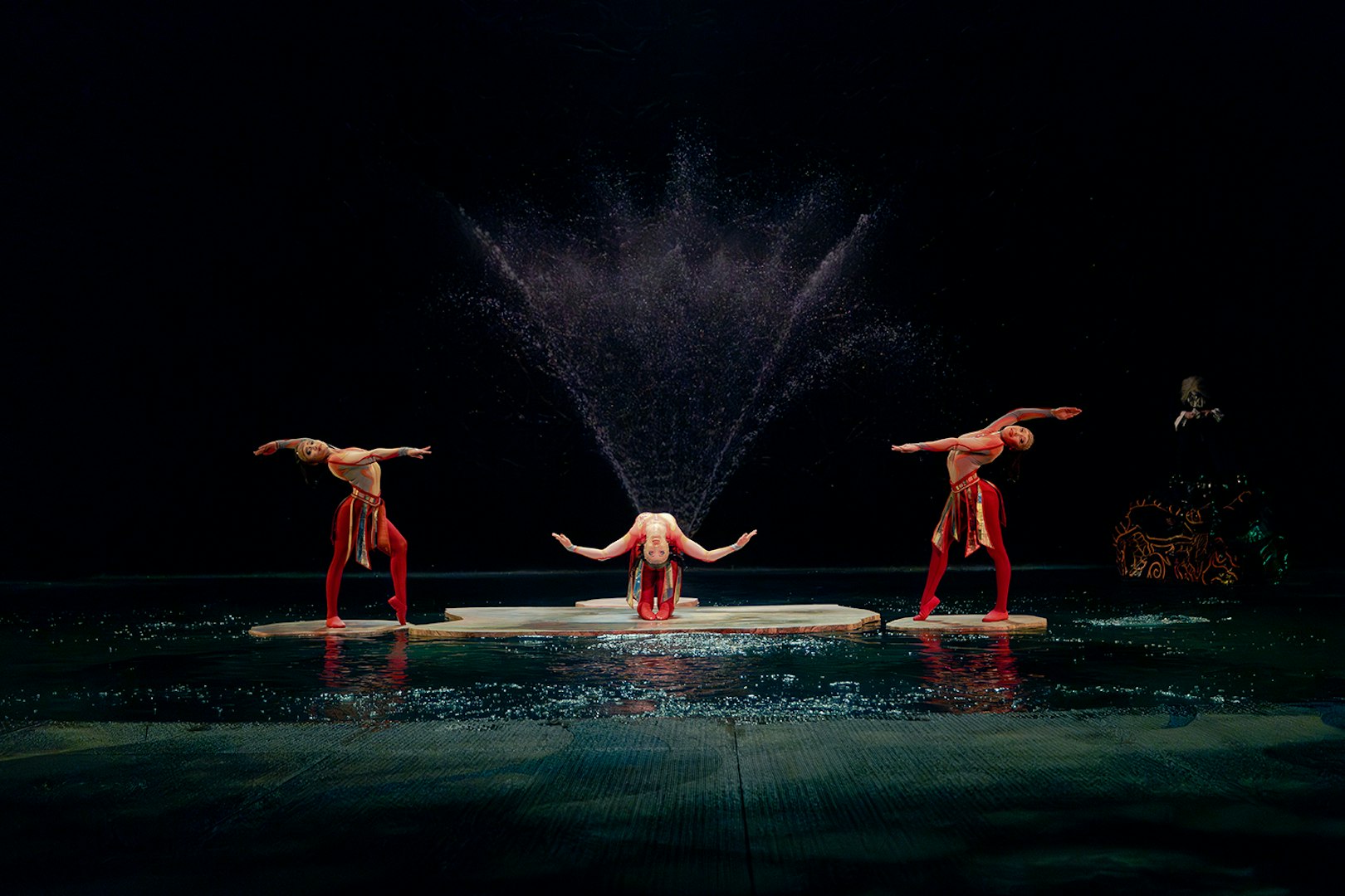 Performers in a water-themed acrobatic show with fountains in the background.