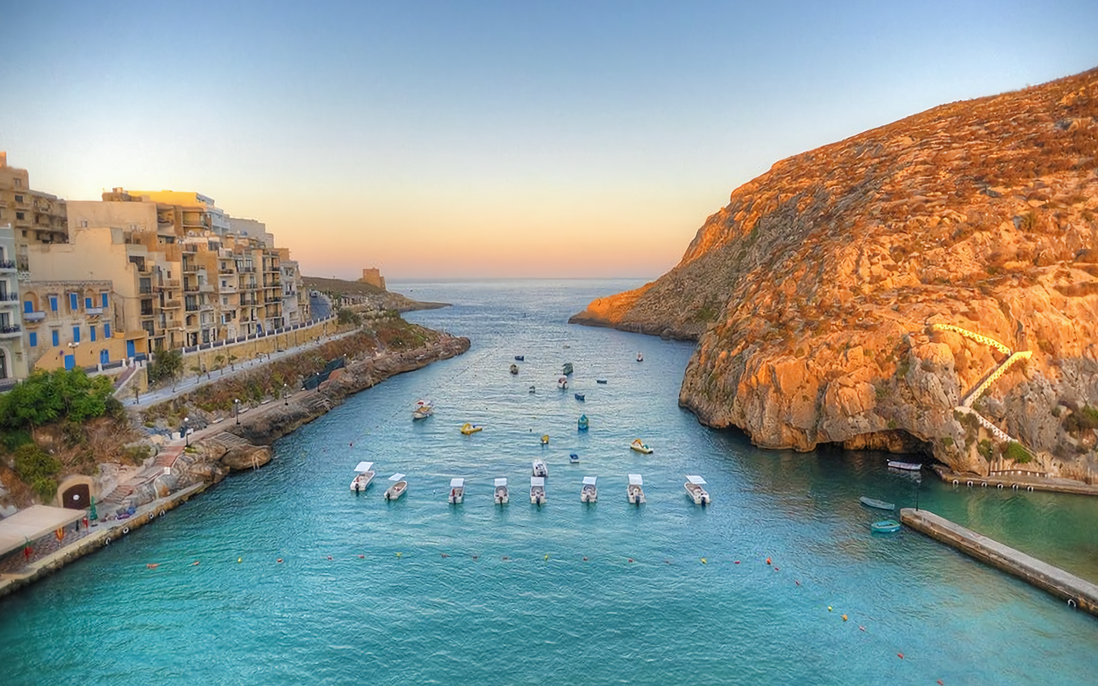Boats in Xlendi Bay, Gozo with cliffs and buildings at sunset.