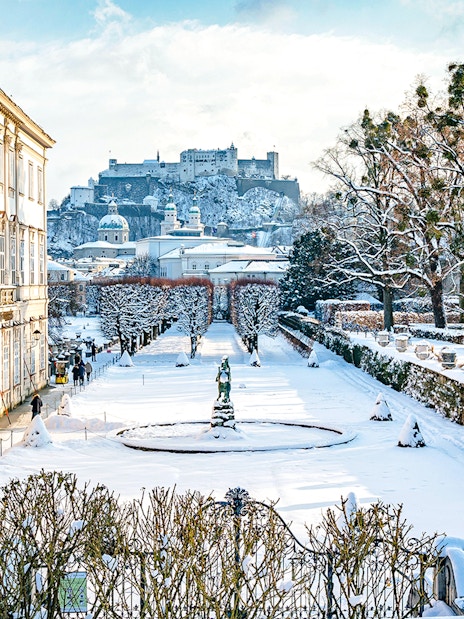 Mirabell Gardens in Salzburg covered in winter snow with Hohensalzburg Fortress in the background.