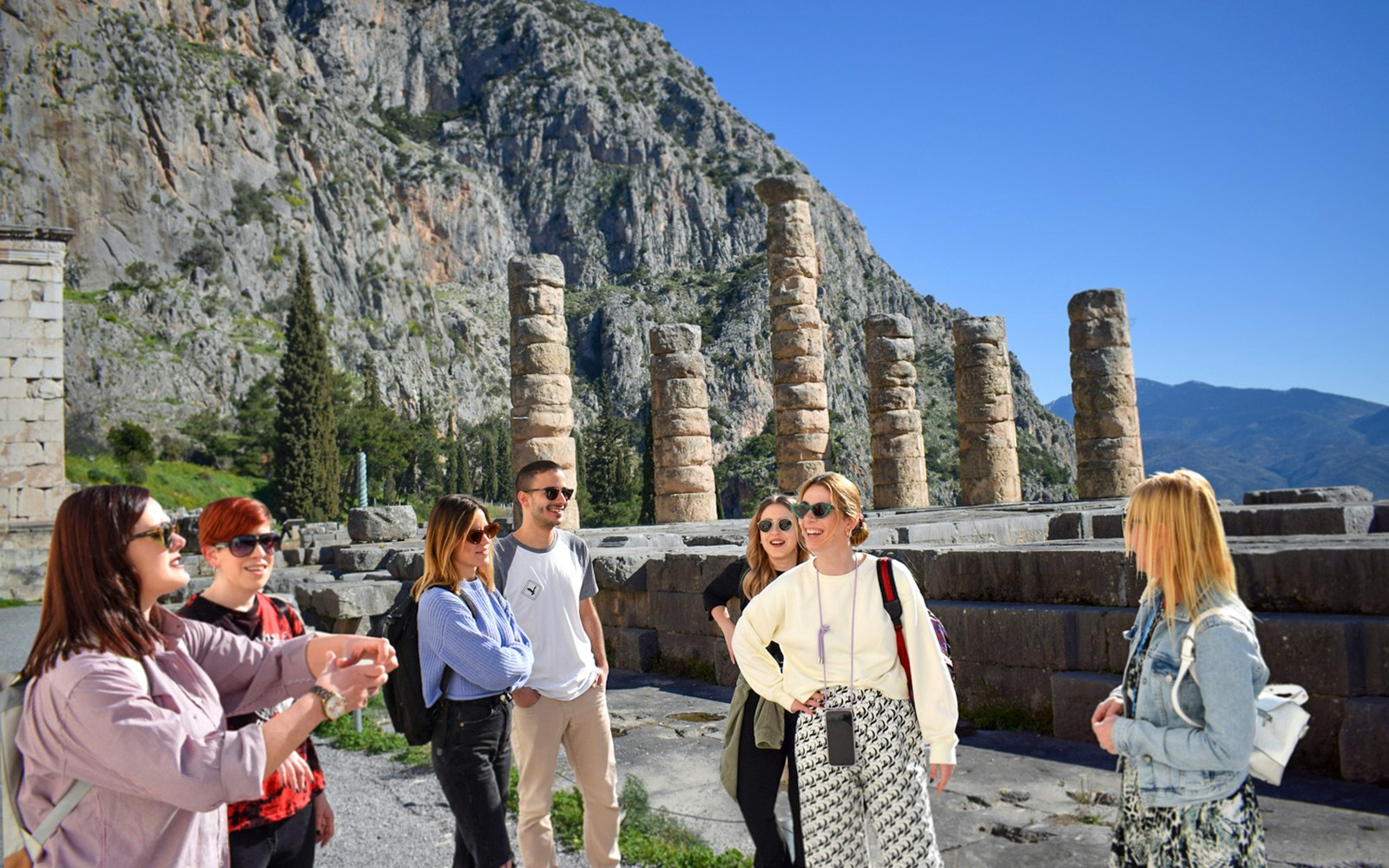 Visitors exploring ancient ruins at Delphi, Greece, with mountainous backdrop.