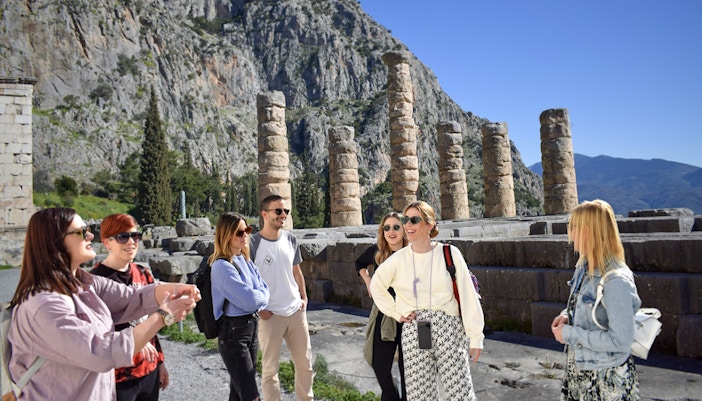 Visitors exploring ancient ruins at Delphi, Greece, with mountainous backdrop.
