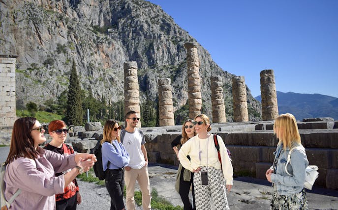 Visitors exploring ancient ruins at Delphi, Greece, with mountainous backdrop.