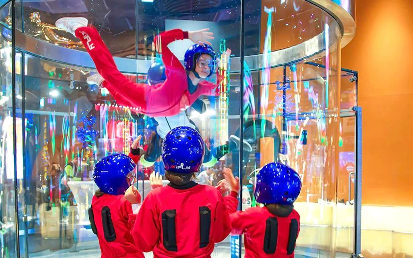 Indoor skydiving experience at iFLY with children watching in flight suits and helmets.
