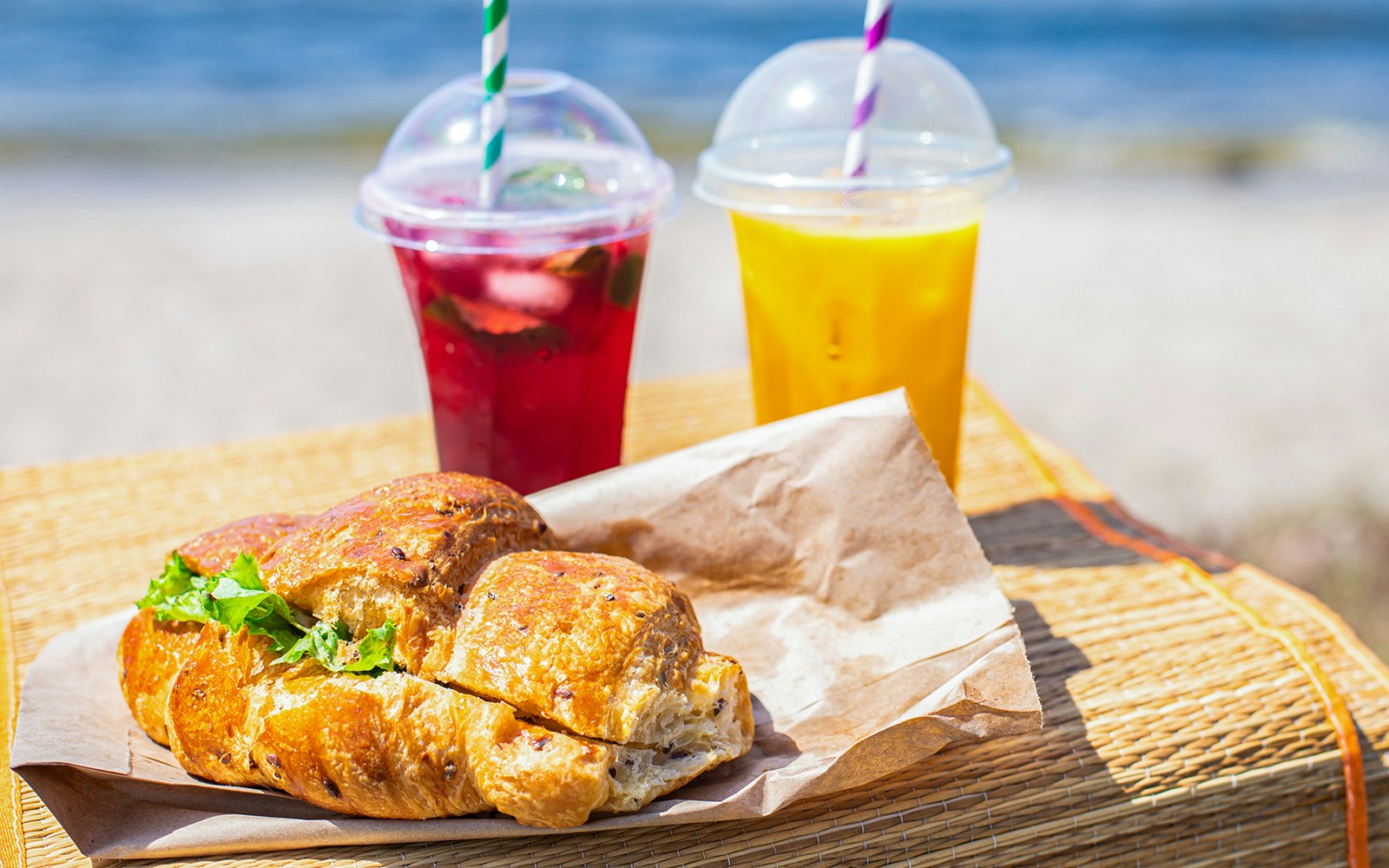 Croissant sandwich and drinks on a beach picnic setup.