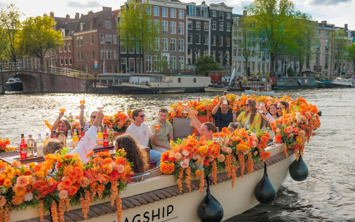 Luxury boat cruise with people enjoying drinks on Amsterdam canal, surrounded by orange flowers.