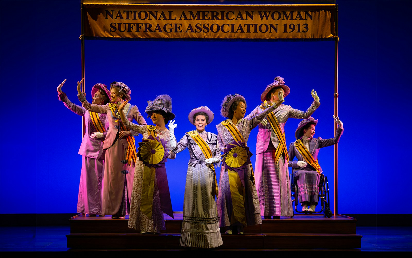 Women in period costumes with sashes on stage under "National American Woman Suffrage Association 1913" banner.