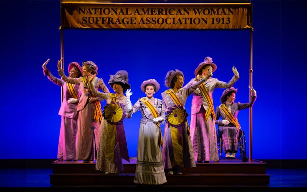 Women in period costumes with sashes on stage under "National American Woman Suffrage Association 1913" banner.