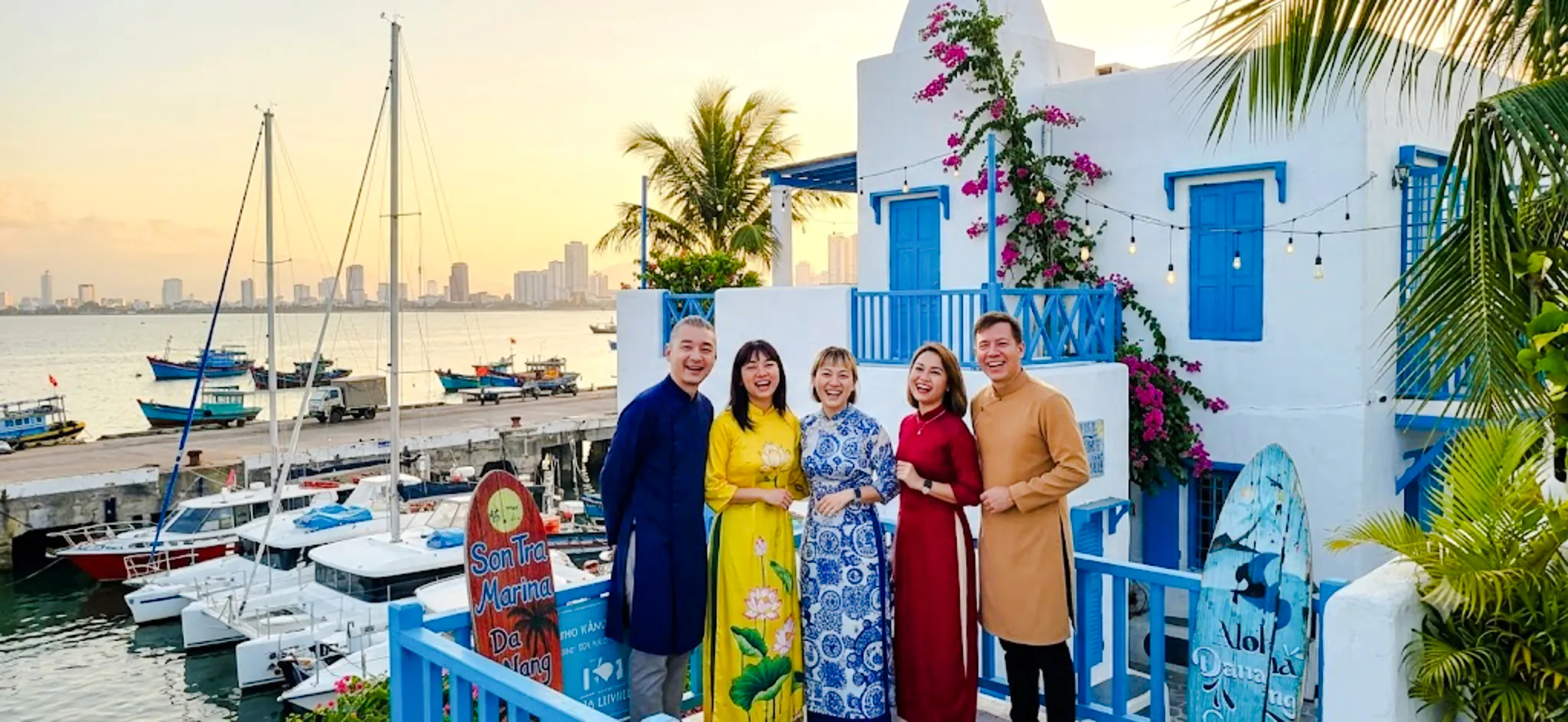 Group enjoying sunset at Son Tra Marina, Da Nang, with colorful buildings and boats in the background.