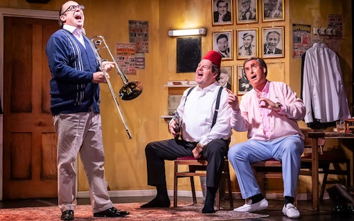 Three men in a comedic scene, one playing a trombone, in a room with vintage posters and photos.