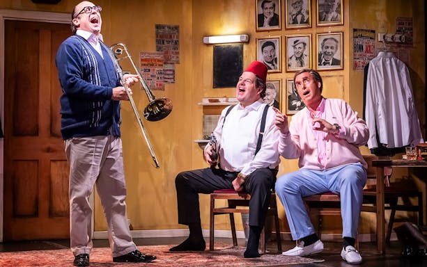 Three men in a comedic scene, one playing a trombone, in a room with vintage posters and photos.