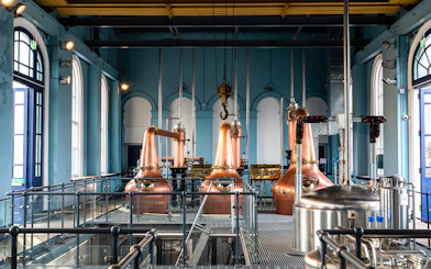 Copper stills inside Titanic Whiskey Distillery with arched windows and industrial equipment.