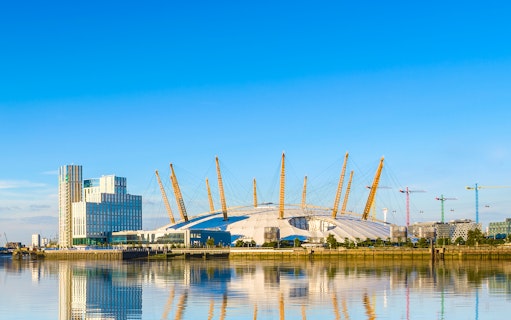 O2 Arena in London with surrounding buildings and cranes reflected in the Thames River.