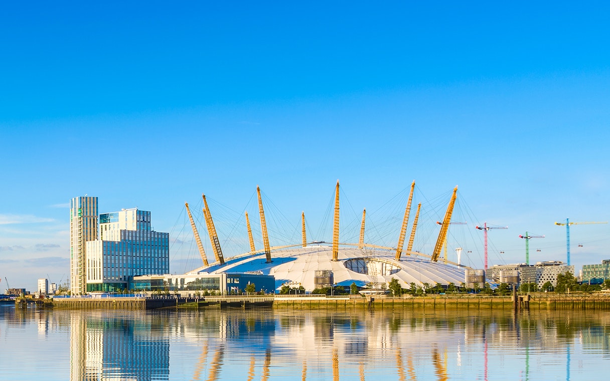 O2 Arena in London with surrounding buildings and cranes reflected in the Thames River.